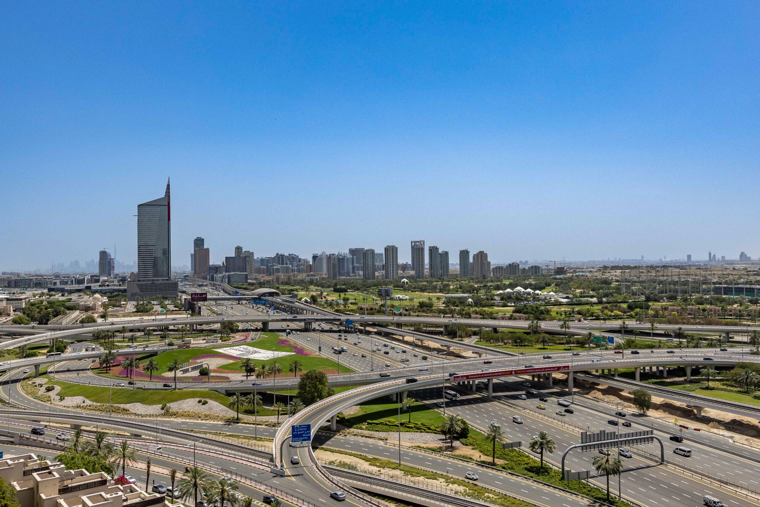 Aerial daytime view of a Dubai highway interchange with a flag-striped spire tower against a clear blue sky