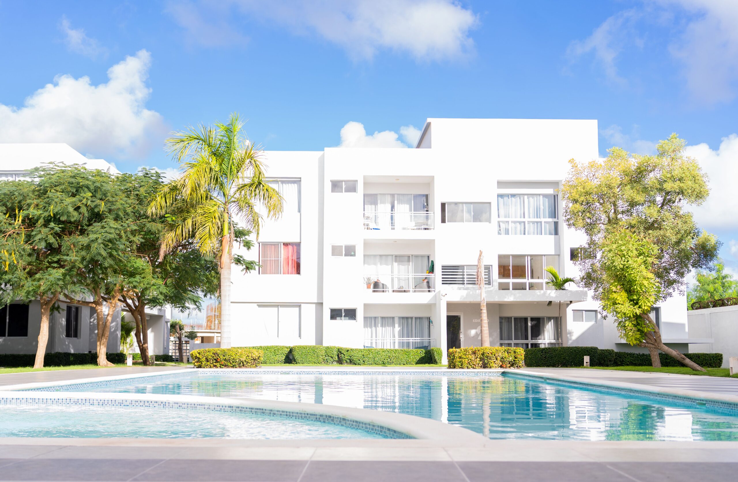 Low-rise white modern apartment building with a surrounding outdoor pool and palm trees under a clear blue sky, representative of resort-style Dubai residential architecture