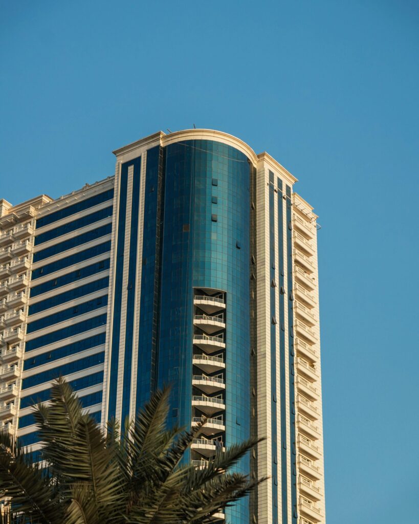 Blue-glass Dubai high-rise apartment tower with a curved balcony feature and palm tree in the foreground under a clear sky
