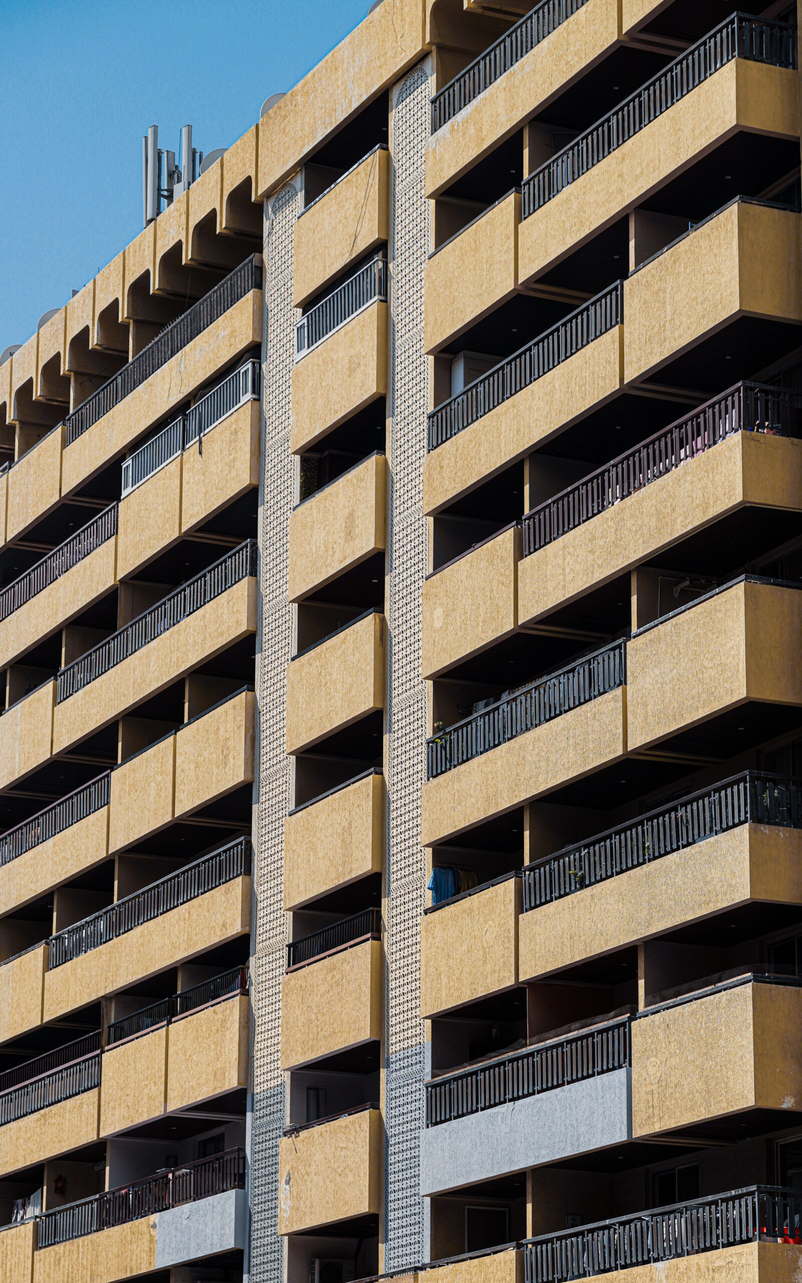 Close-up of a modern tan-toned apartment building facade with dark-railed balconies against a clear blue sky, representative of Candace Aster in Al Furjan, Dubai