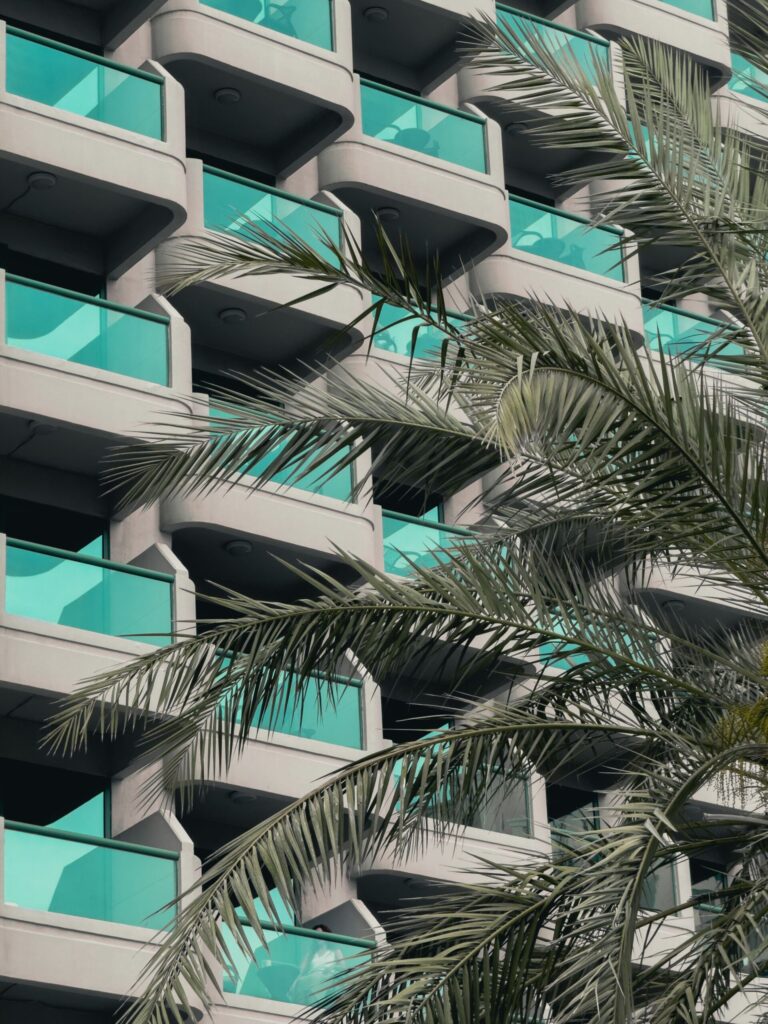 Modern residential apartment building exterior with teal glass balconies and a palm frond in the foreground, Dubai, representative of Joya Blanca, Arjan