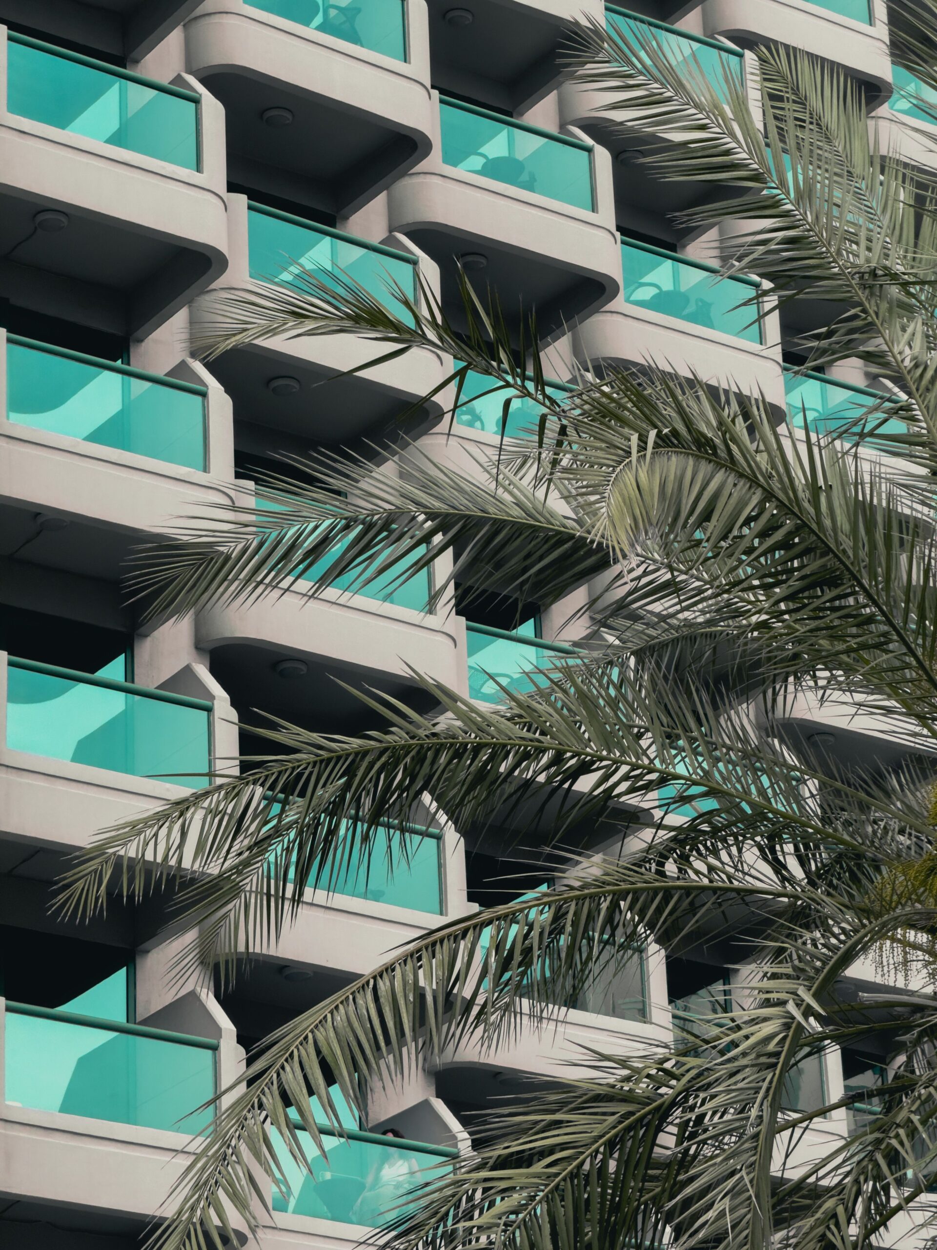 Modern residential apartment building exterior with teal glass balconies and a palm frond in the foreground, Dubai, representative of Joya Blanca, Arjan
