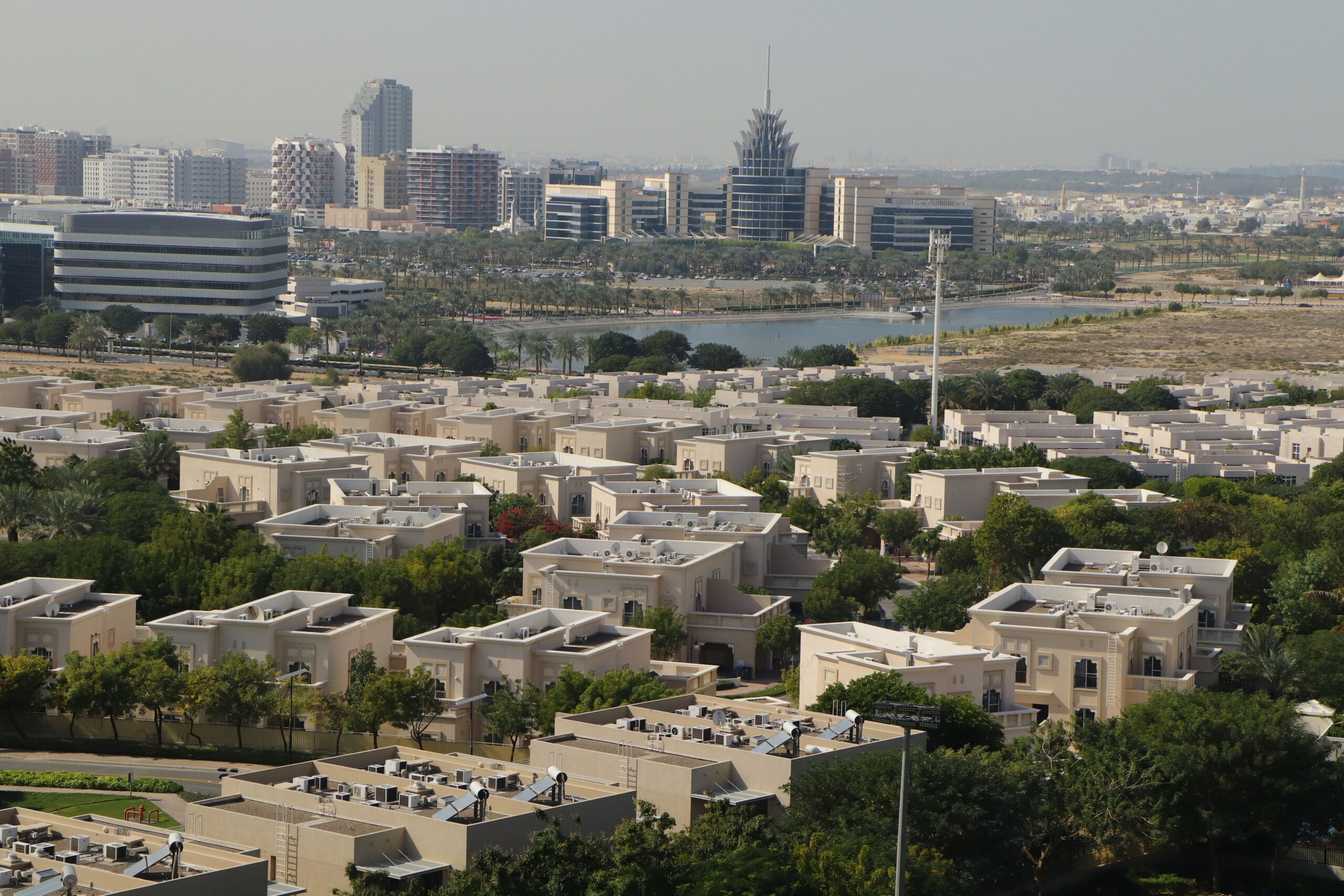 Aerial view of a Dubai villa community with a central lake, cream low-rise villa rooftops in the foreground and Dubai's Internet City and Media City skyline visible in the distance, representative of Springs 6, The Springs, Emirates Living