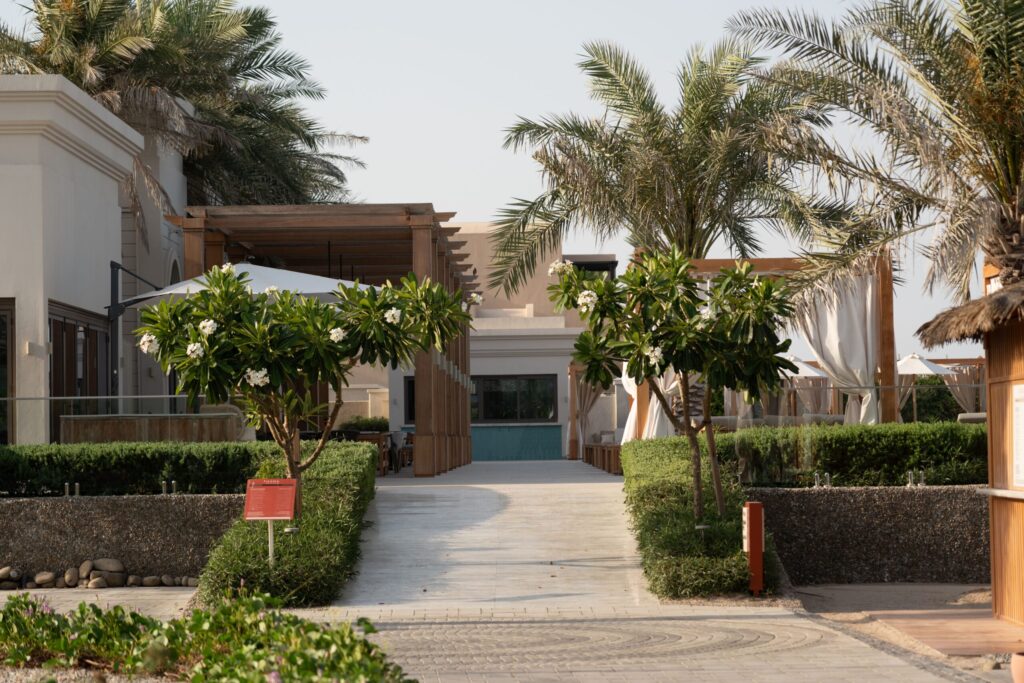 Palm-lined walkway between low-rise beige buildings with a wooden pergola and pool in a Dubai residential community, no people visible