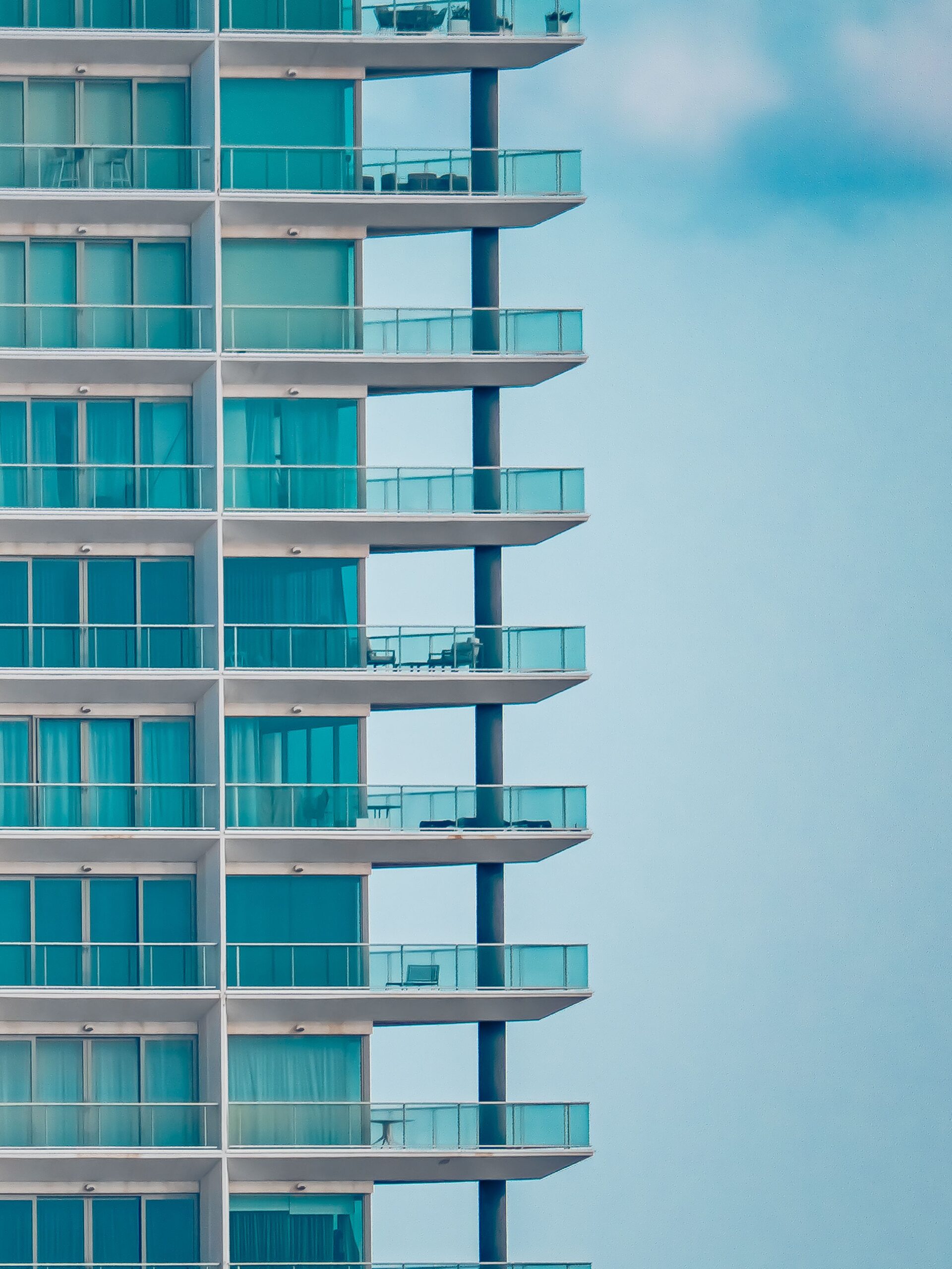 Close-up of a modern glass high-rise apartment tower facade with teal-tinted balconies and outdoor furniture under a clear sky, representative of contemporary waterfront residential architecture in Dubai Creek Harbour where The Grand at Dubai Creek Harbour is located