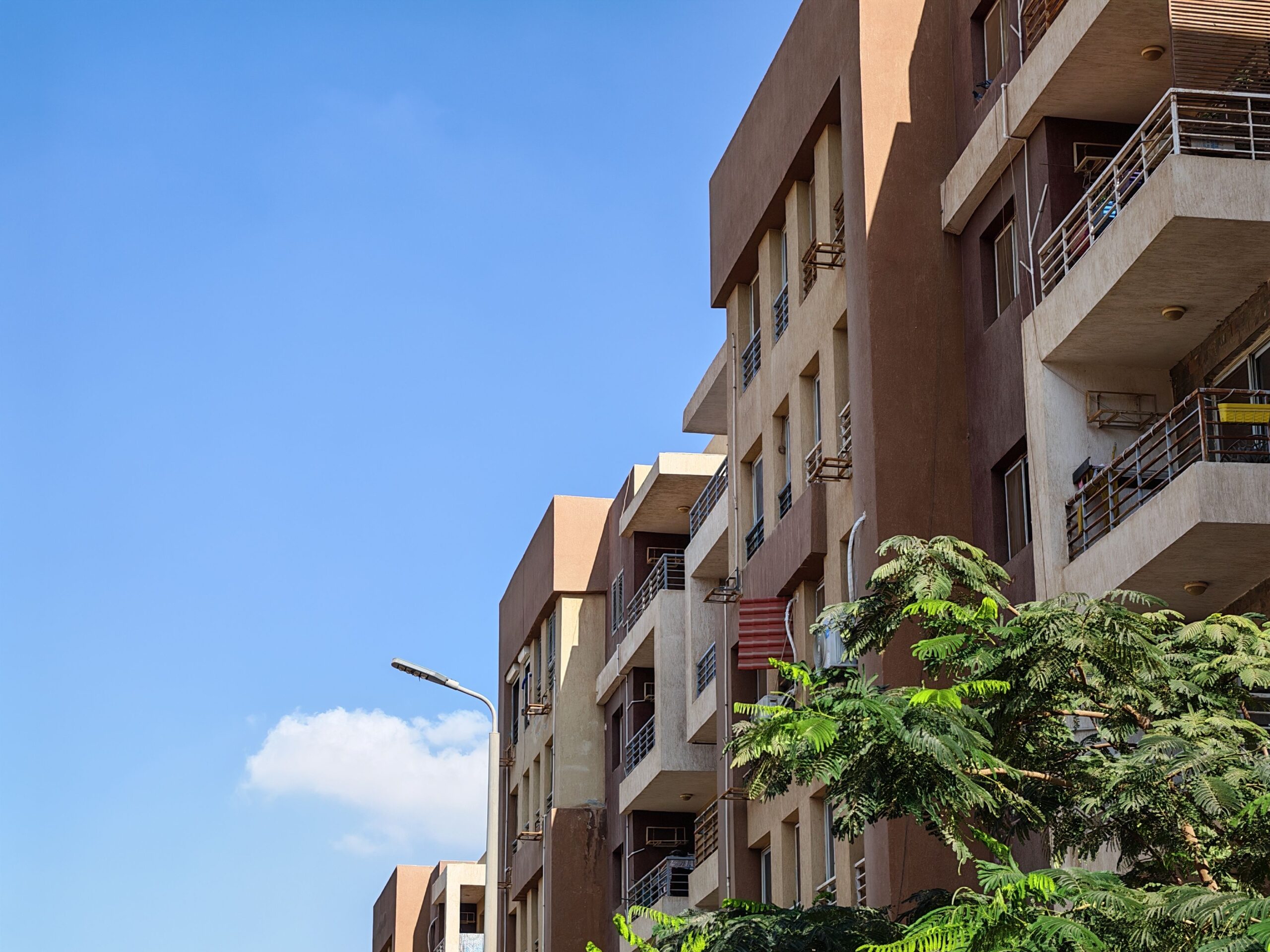 Low-rise beige apartment building with balconies and greenery under a clear blue sky, similar in style to Park Lane by Heilbronn in JVC