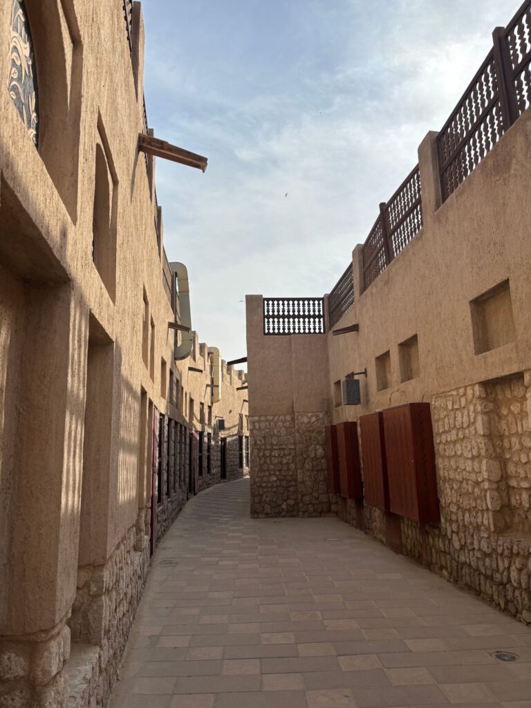 Empty sandstone Old Town-style heritage alley with arched stonework and dark wood-lattice balcony screens in Downtown Dubai, near Reehan 8