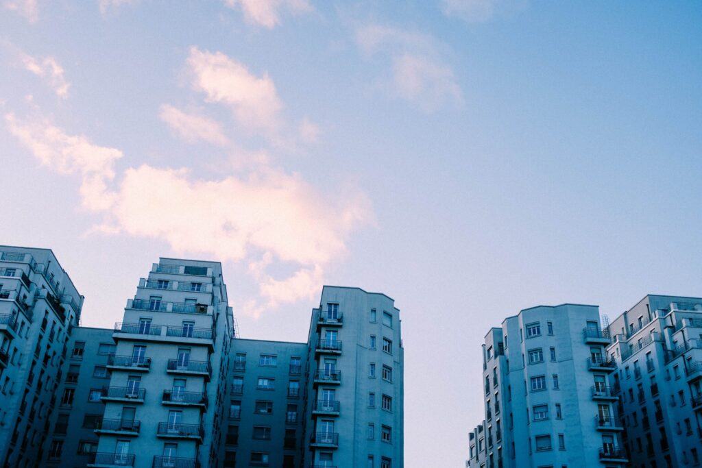 Pale blue apartment buildings with balconies rising toward a soft pink and blue sky