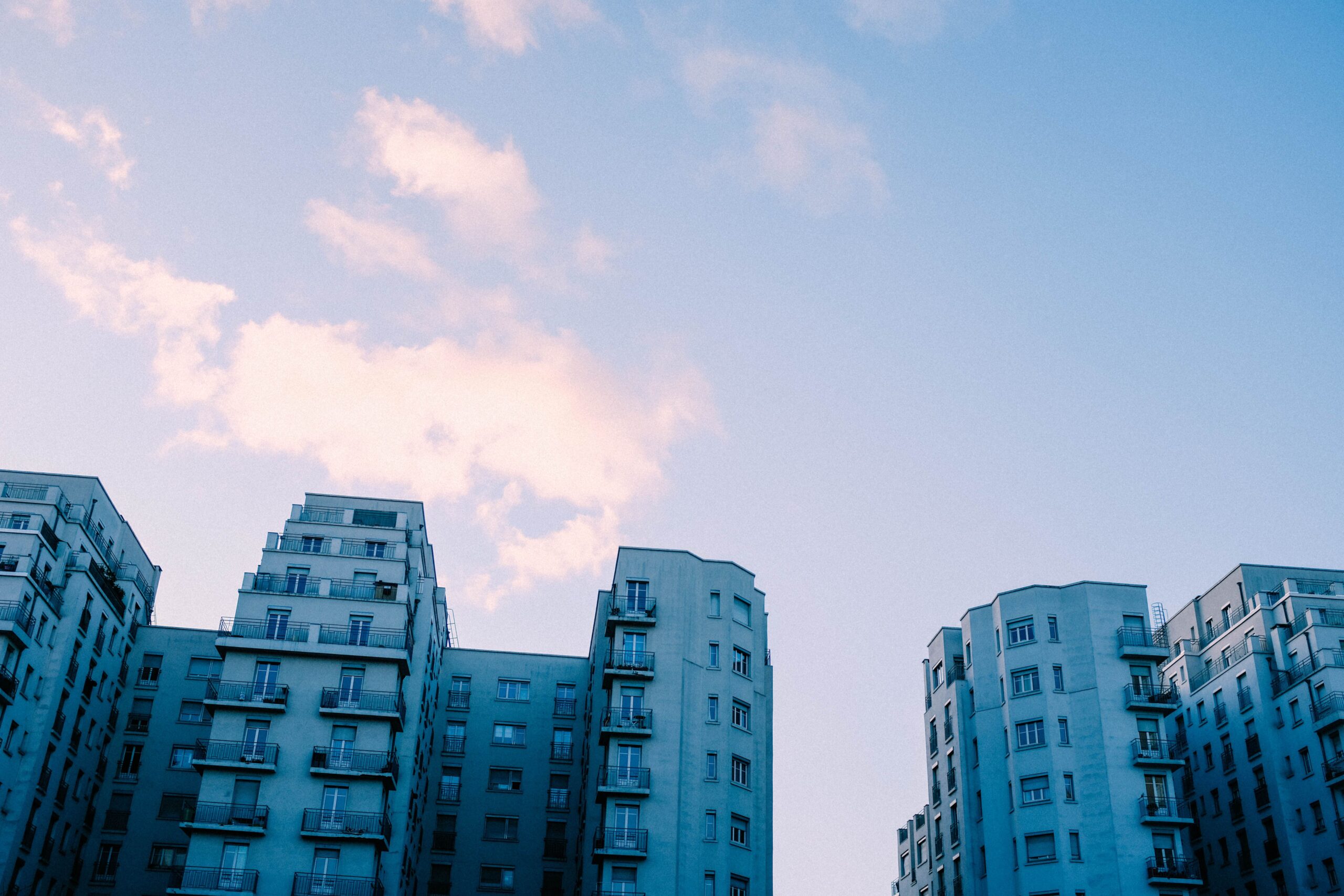 Pale blue apartment buildings with balconies rising toward a soft pink and blue sky