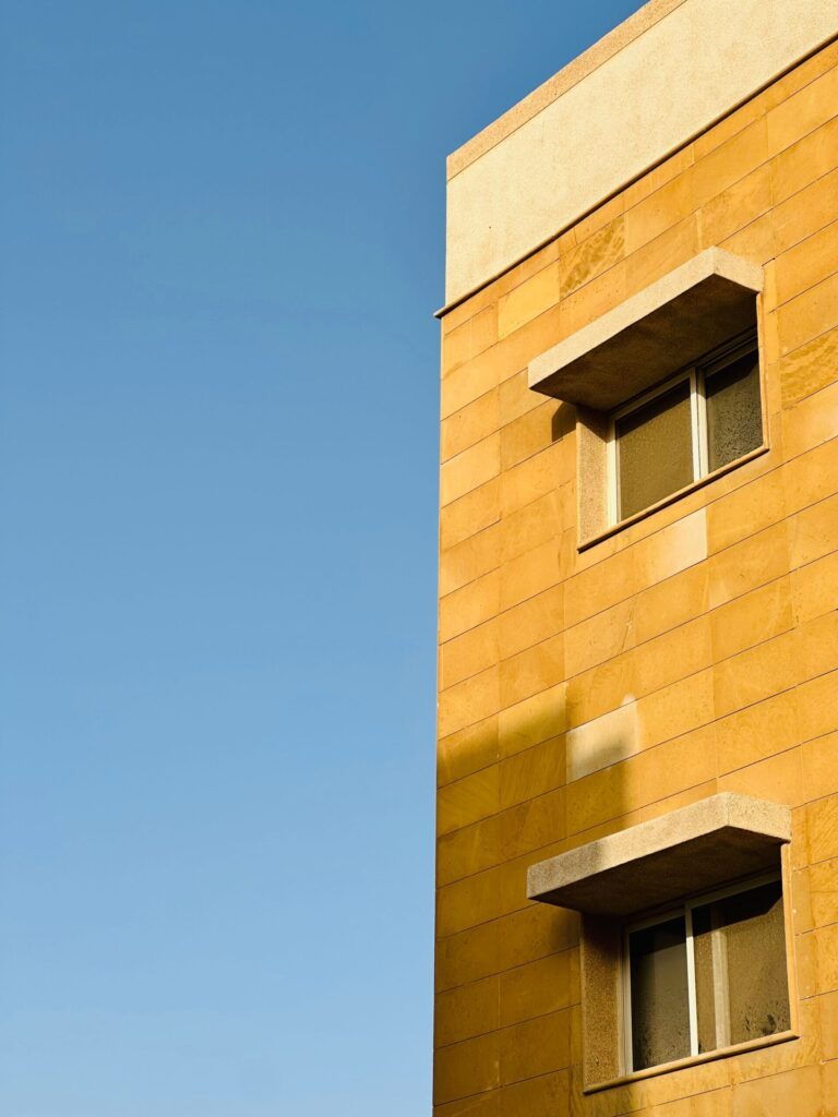 Sandstone-toned low-rise residential building facade against a clear blue sky, similar in style to Golden Wood Views, JVT