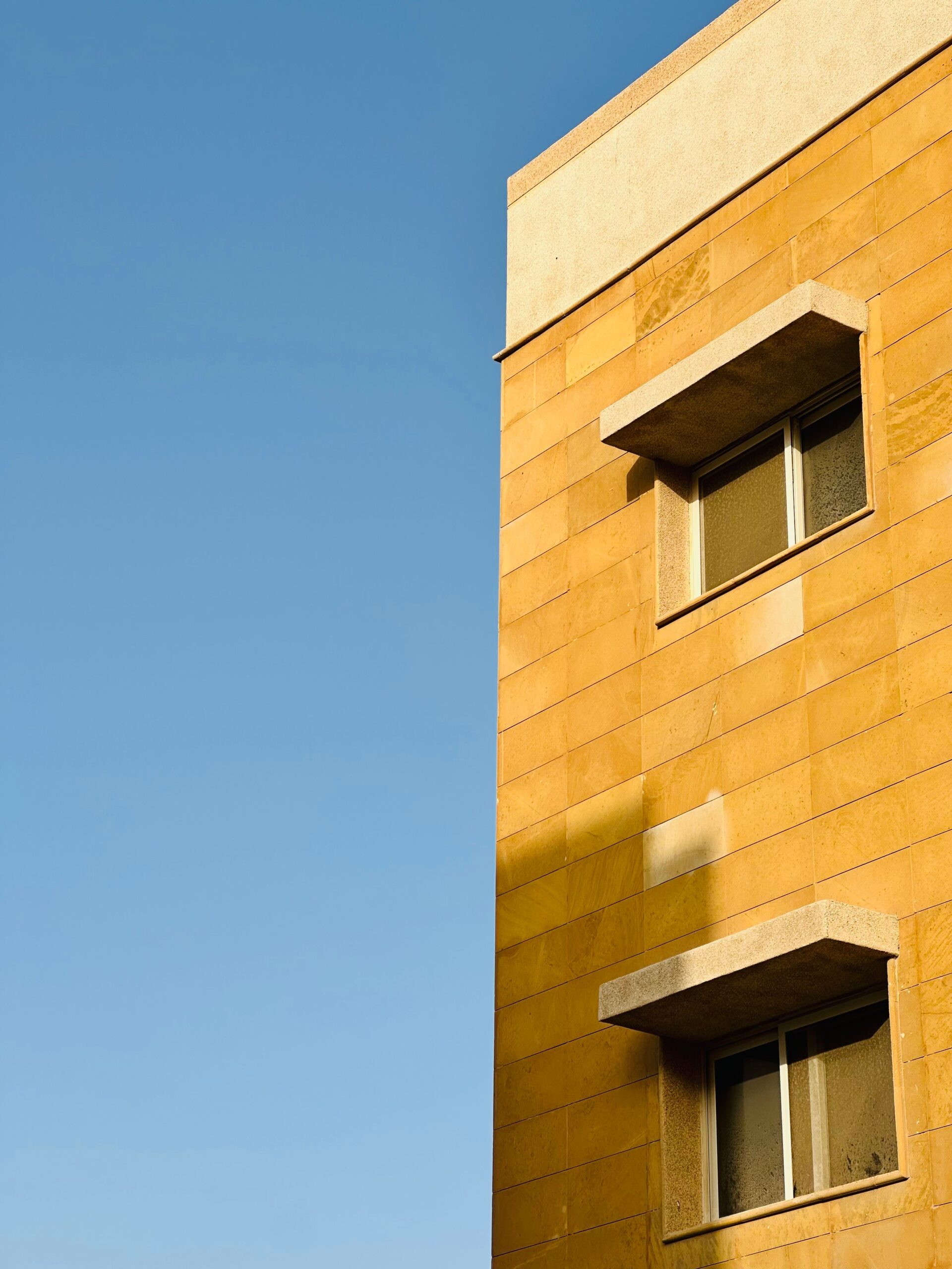 Sandstone-toned low-rise residential building facade against a clear blue sky, similar in style to Golden Wood Views, JVT