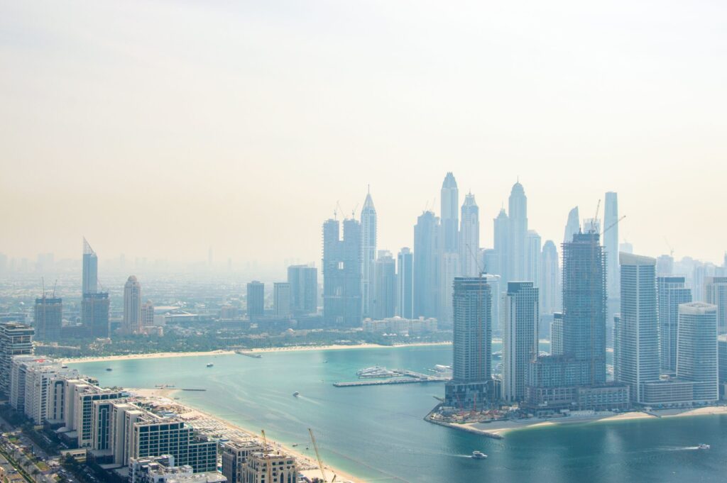 Hazy daytime aerial view of a Dubai waterfront skyline with high-rise residential towers along a lagoon-style coastline, illustrative of the Meydan waterfront setting of Azizi Riviera 69