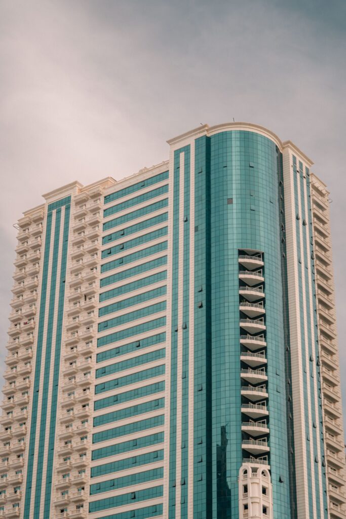 Modern high-rise residential tower with teal glass and stone facade under a pale blue sky in Dubai
