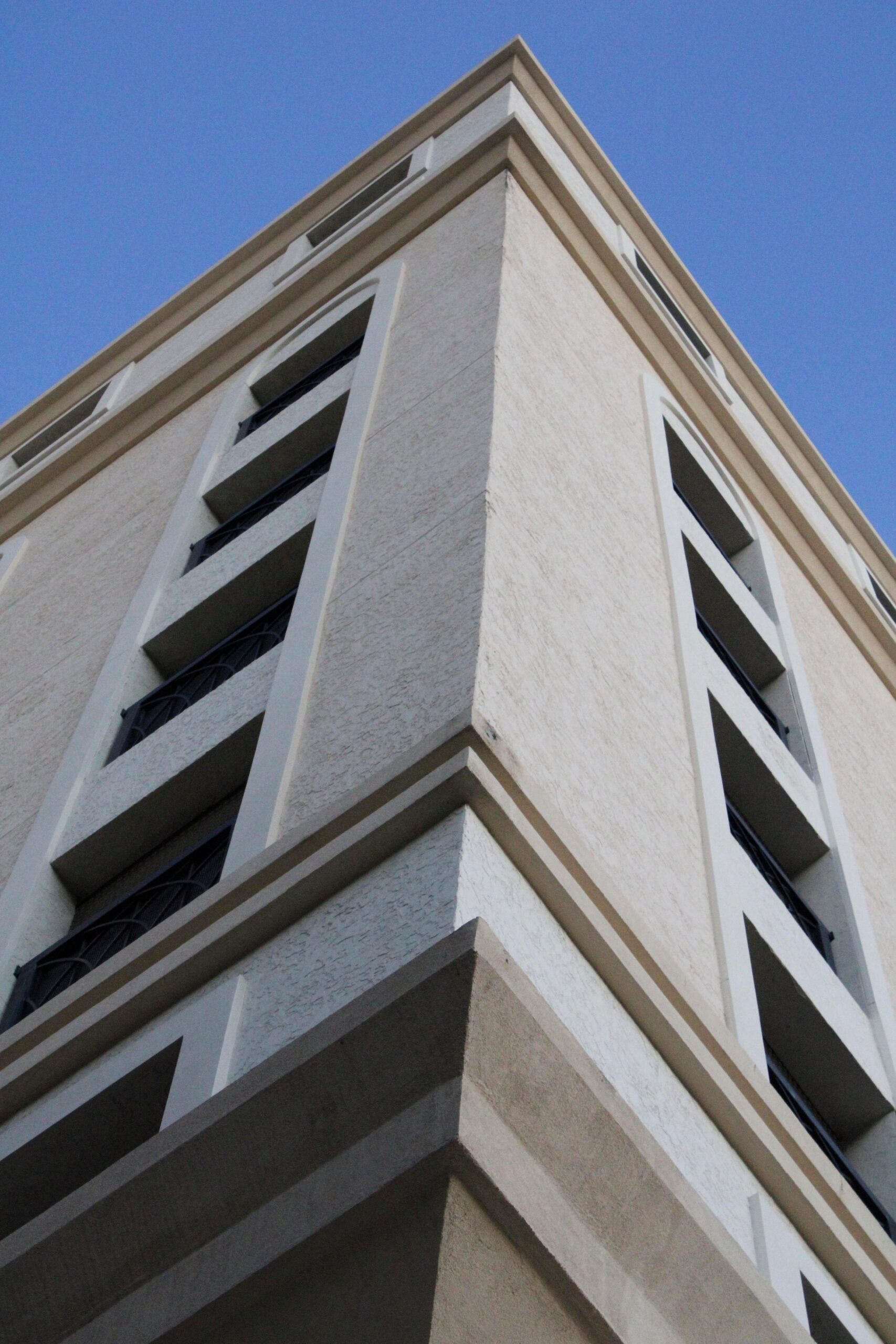 Upward view of a cream-toned modern residential building corner with arched balcony openings against a clear blue sky, representative of Gardenia Livings, Arjan