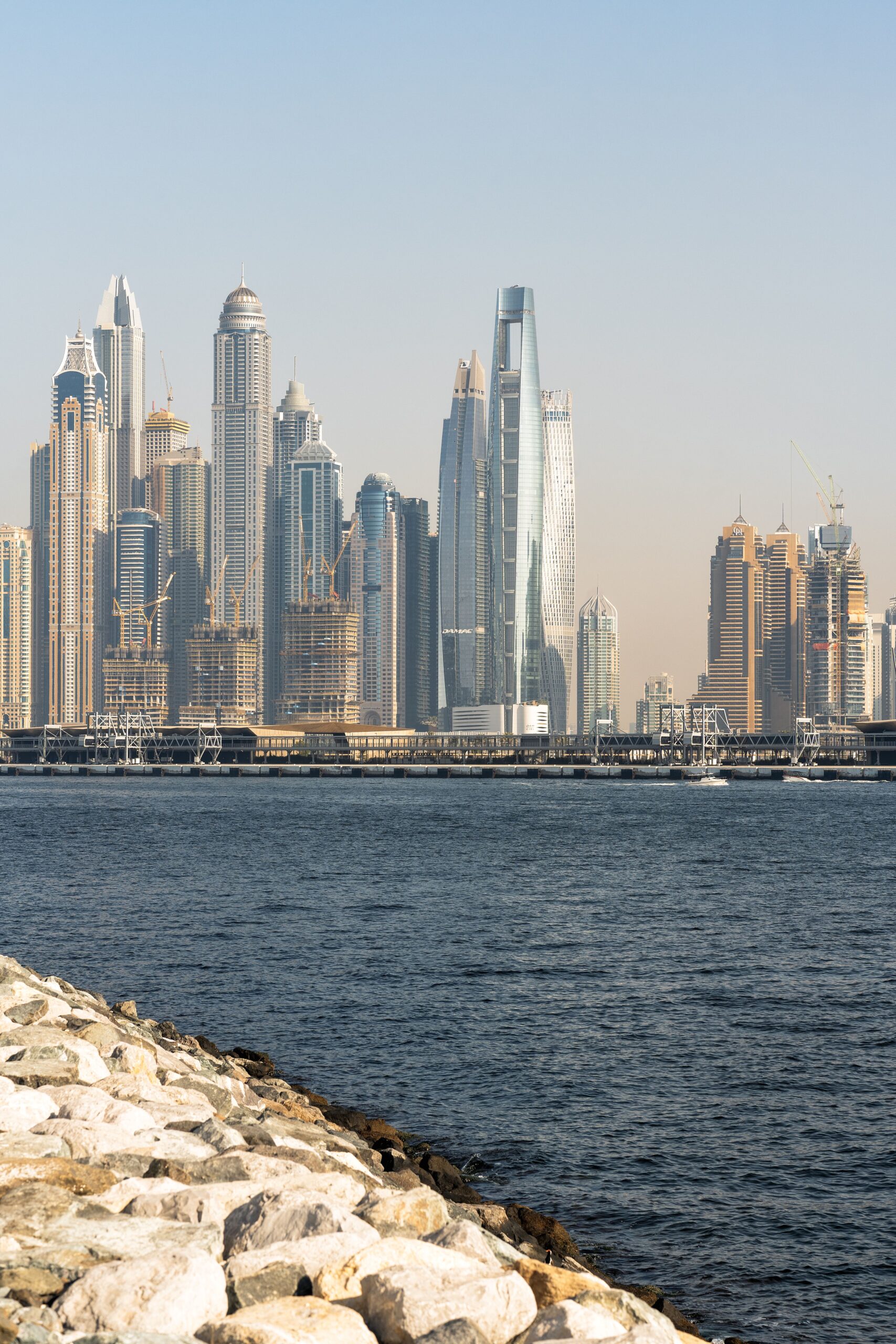 Daytime Dubai skyline with modern towers under construction, seen across the water from a rocky waterfront