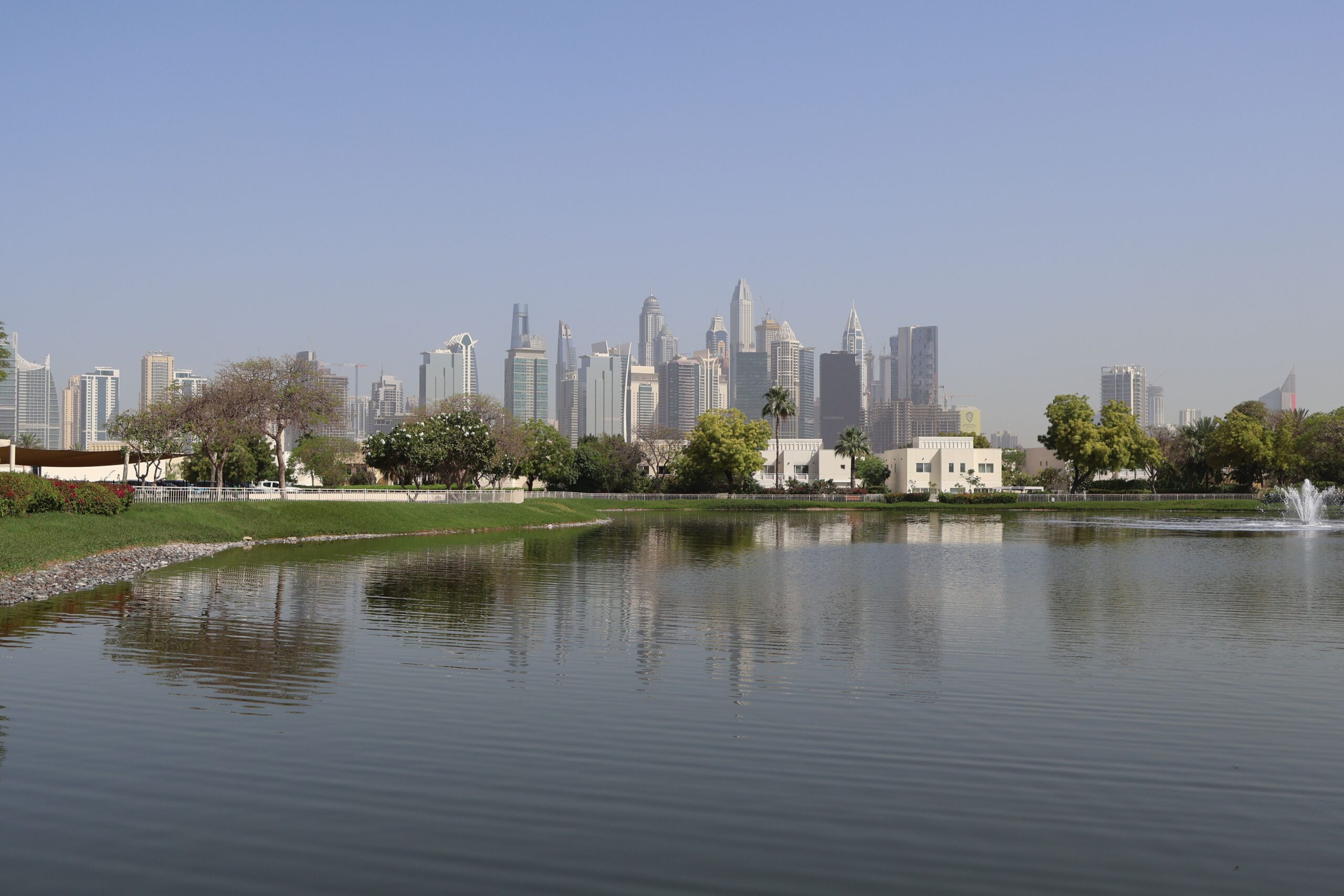 Daytime view of a landscaped park lake with a Dubai high-rise skyline in the background, evoking the green, park-adjacent setting of Parkland in Dubai Hills Estate