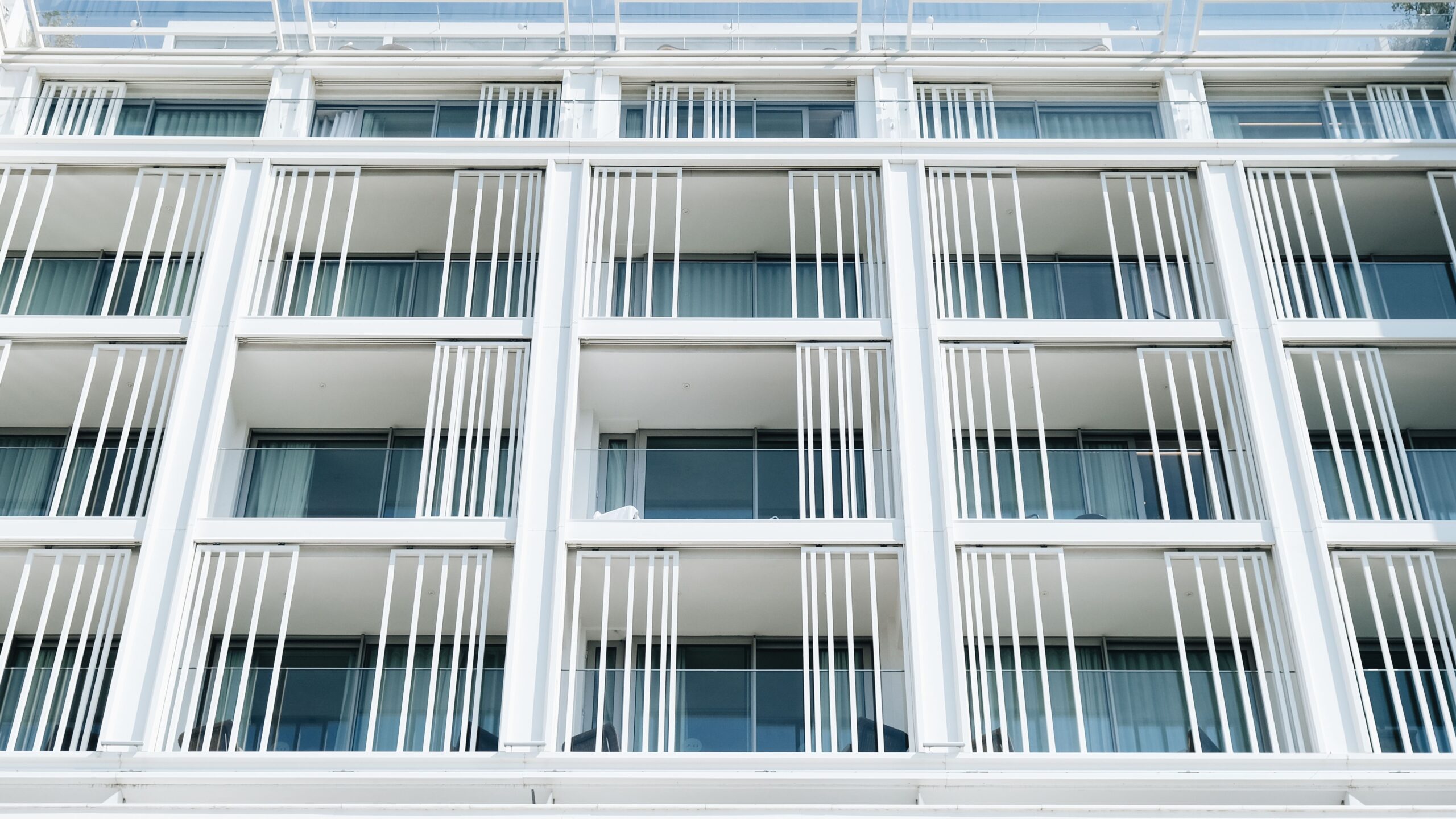 Close-up of a modern white apartment building facade with vertical louvre balcony screens and blue-tinted glass windows, evocative of a Liwan, Dubai residential development