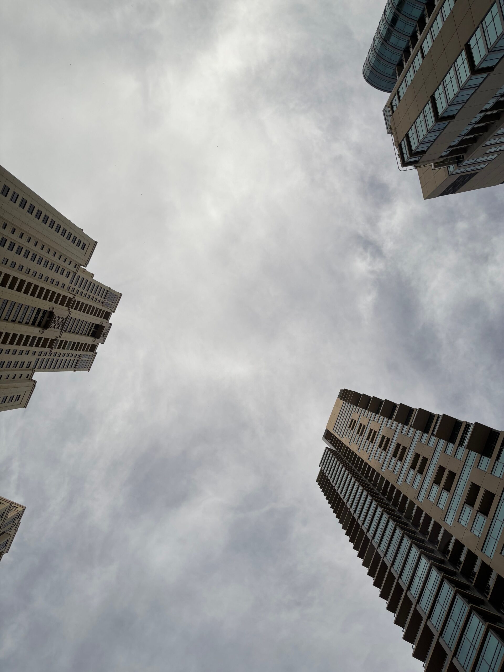 Extreme low-angle view looking up at several modern beige and tan Dubai residential towers with balconies against an overcast sky, representative of the Dubai Production City area near The Towers by OBS