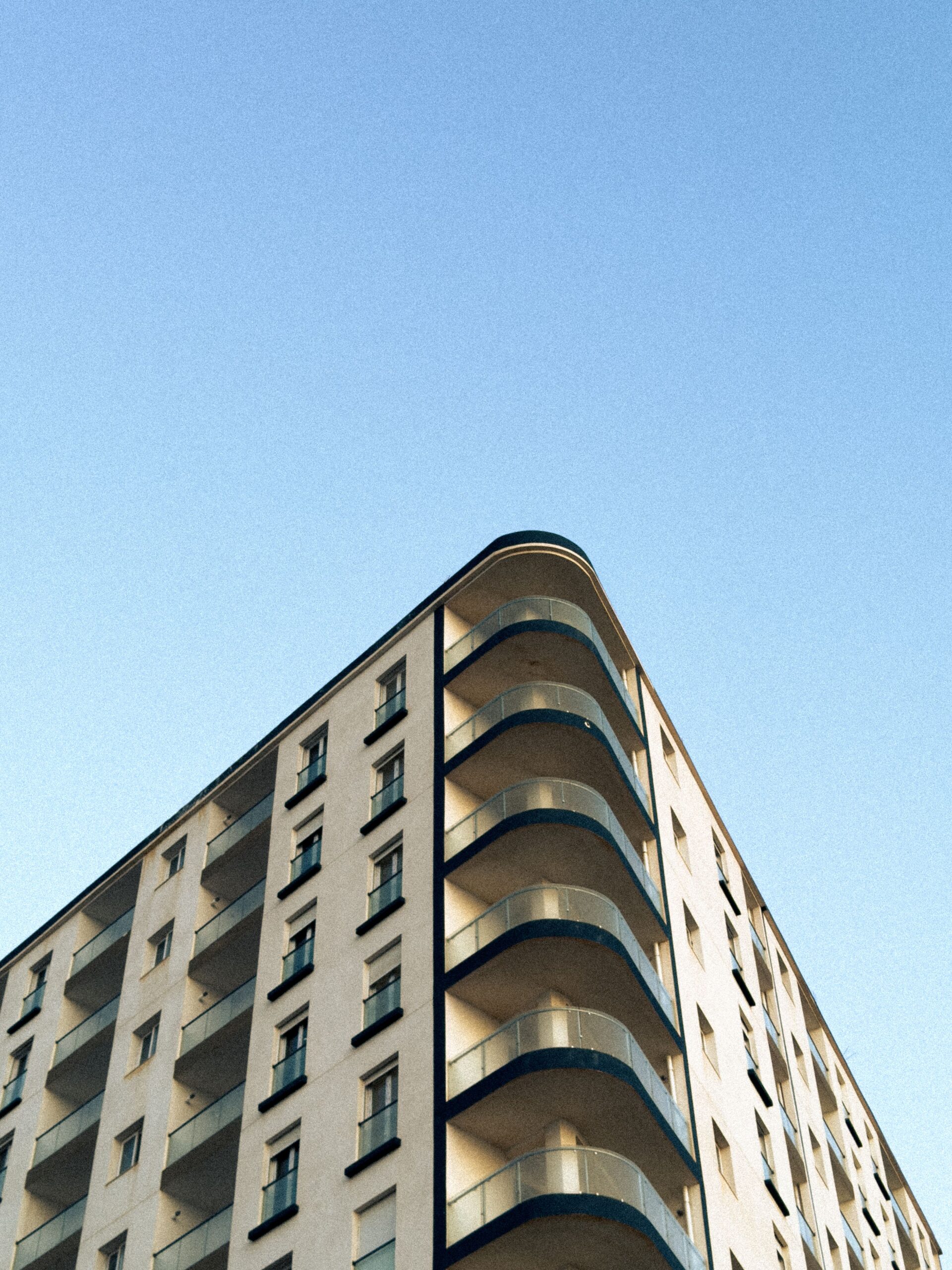 Upward view of a modern cream-toned mid-rise residential building corner with curved balconies against a clear blue sky, representative of Floarea Residence, Arjan