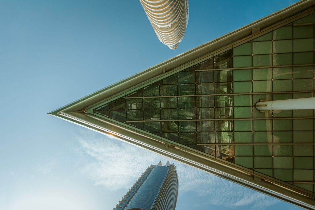 Low-angle view of a green glass high-rise tower corner against a clear blue sky with other Dubai towers in the background, representative of the Jumeirah Lake Towers district near Madina Tower