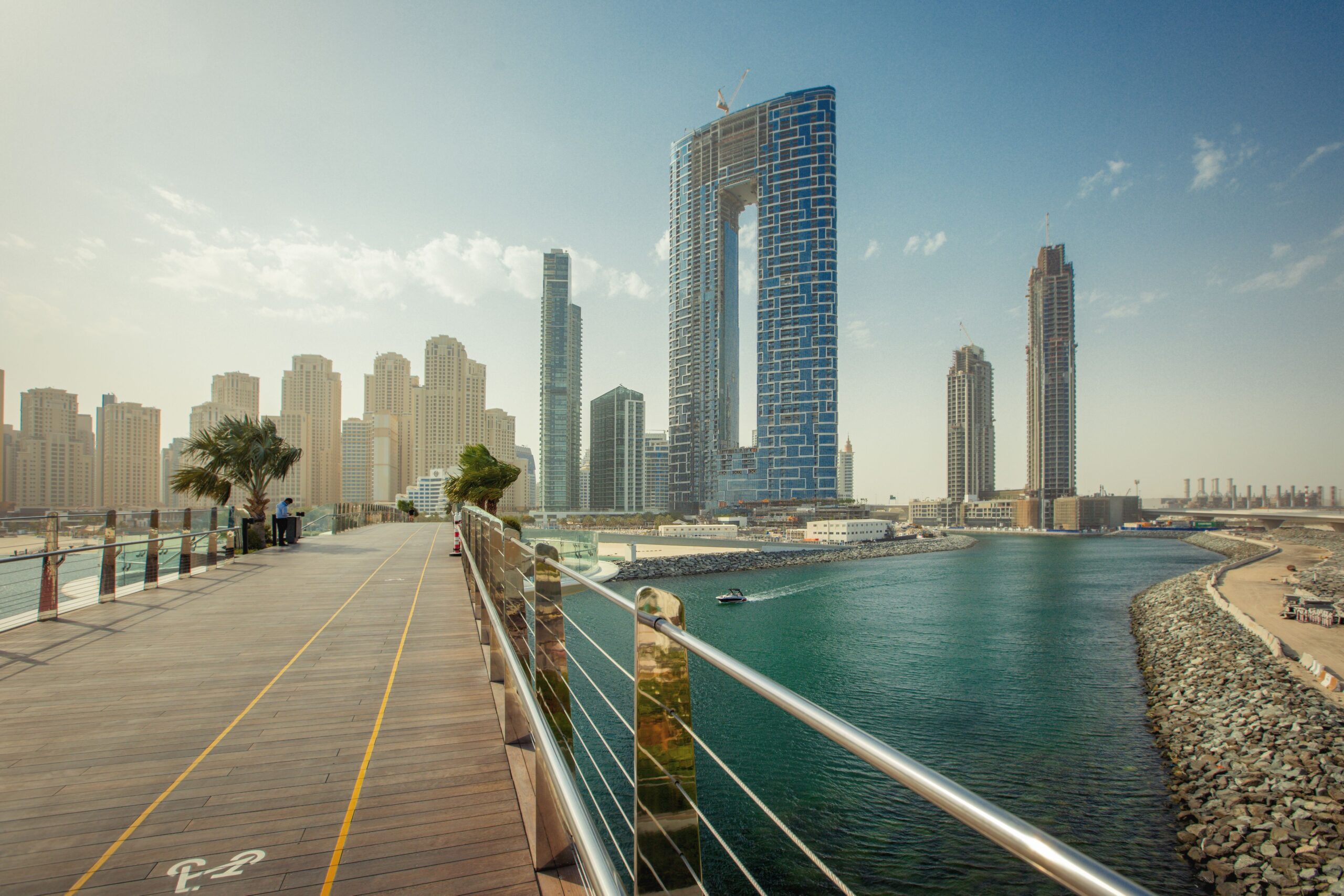 Daytime view of a Dubai waterfront promenade with a modern high-rise tower and clear blue sky