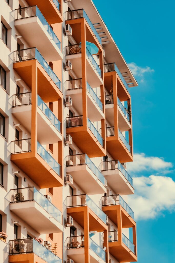 Low angle view of a modern condominium facade with glass balconies and orange and white accents under a clear blue sky, similar in style to Binghatti Gems, JVC