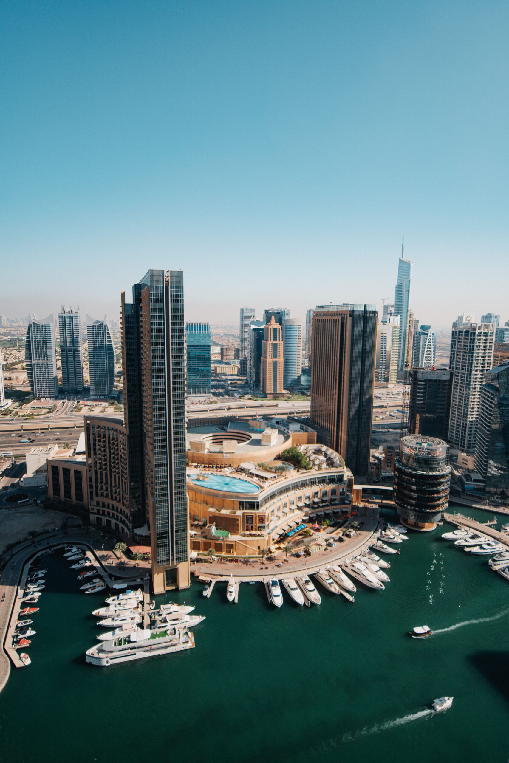 Aerial daytime view of Dubai Marina towers, a rooftop pool deck and a yacht-lined marina waterway