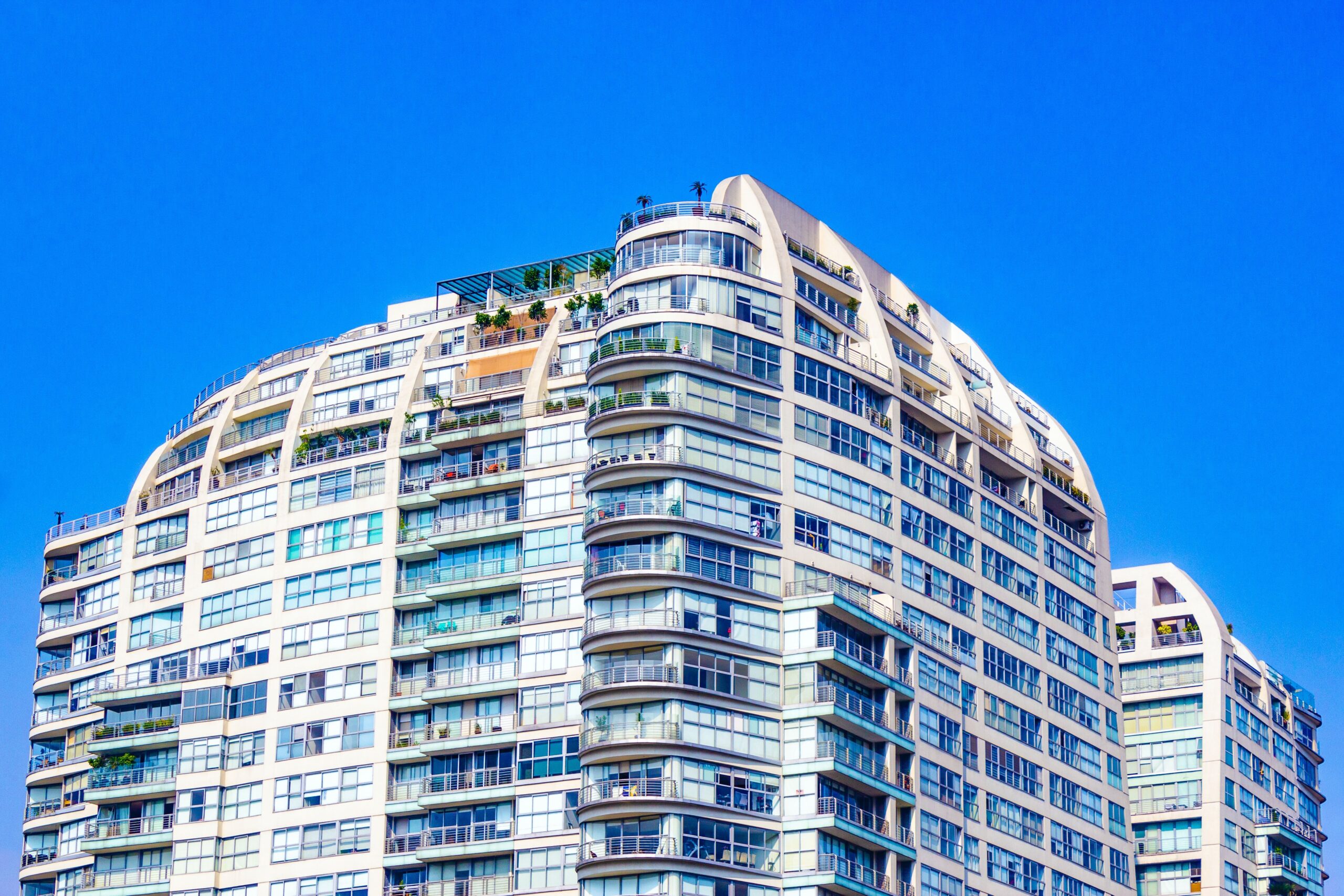 Low angle view of a curved cream modern apartment building corner with a landscaped rooftop terrace against a clear blue sky in Dubai