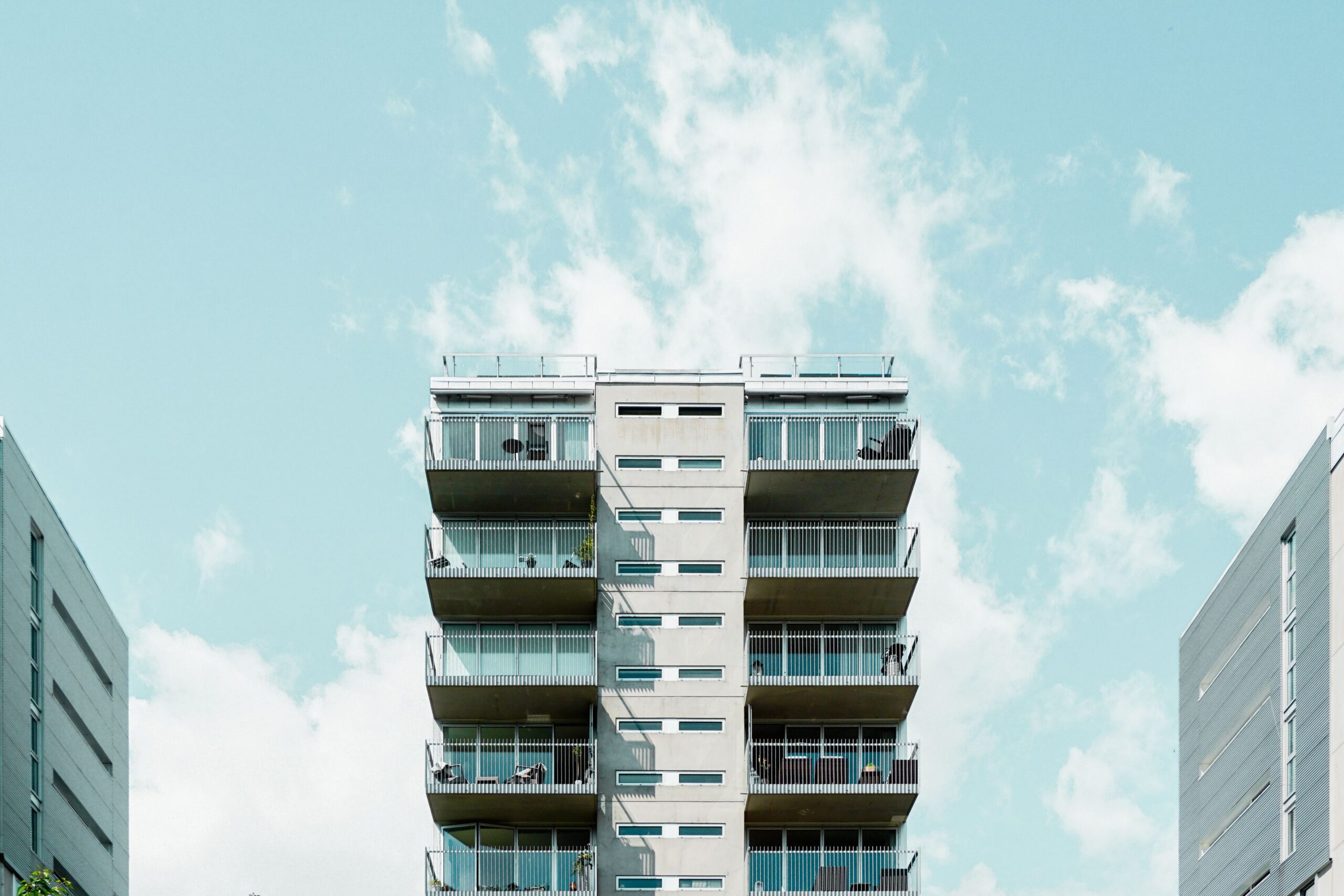 Mid-rise apartment building with balconies rising between two facades under a pale blue sky