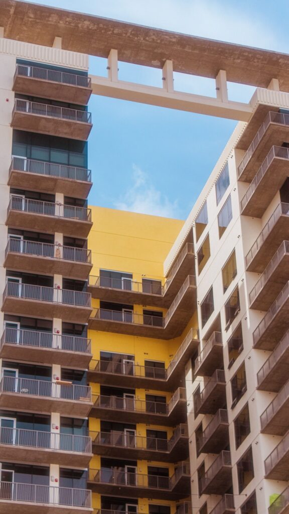 Two connected cream and mustard-yellow modern apartment buildings with balconies linked by an overhead bridge structure under a clear blue sky, representative of contemporary residential architecture near Berton by Azizi in Al Furjan, Dubai