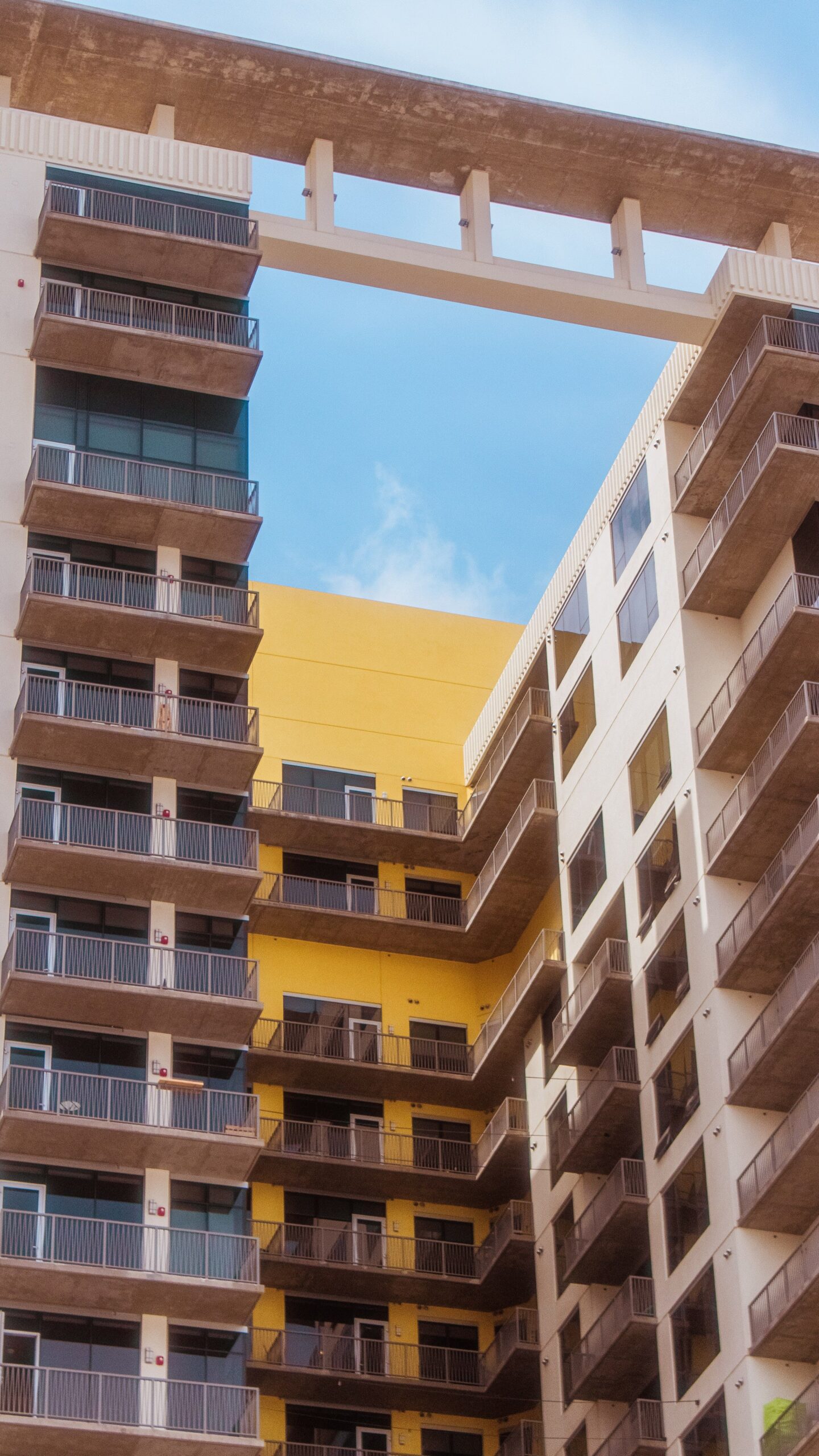 Two connected cream and mustard-yellow modern apartment buildings with balconies linked by an overhead bridge structure under a clear blue sky, representative of contemporary residential architecture near Berton by Azizi in Al Furjan, Dubai