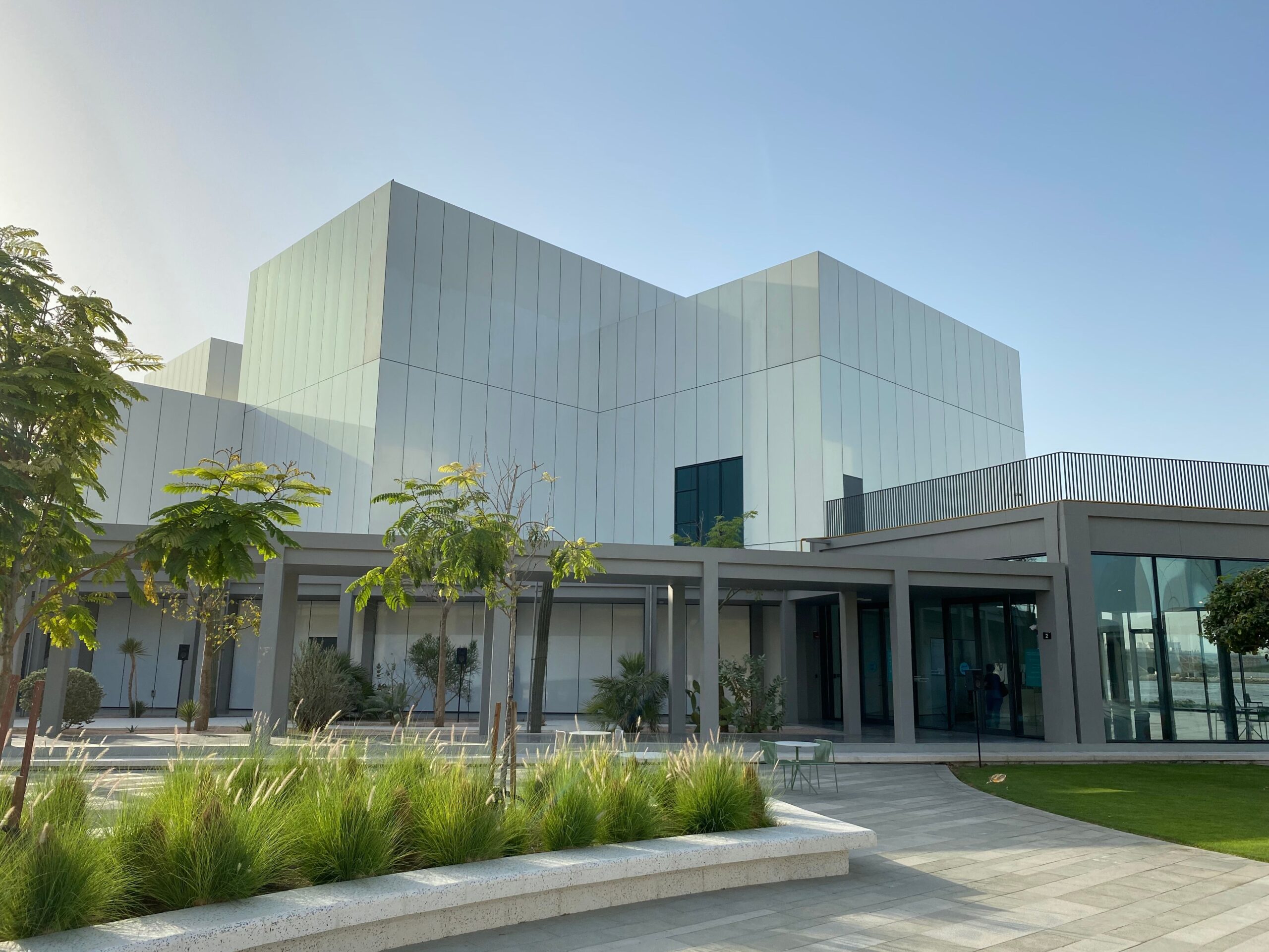 Modern low-rise residential building with a landscaped garden, palm trees and paved walkway under a clear sky, representative of Gardenia Residence's garden-podium setting in Dubai Hills Estate
