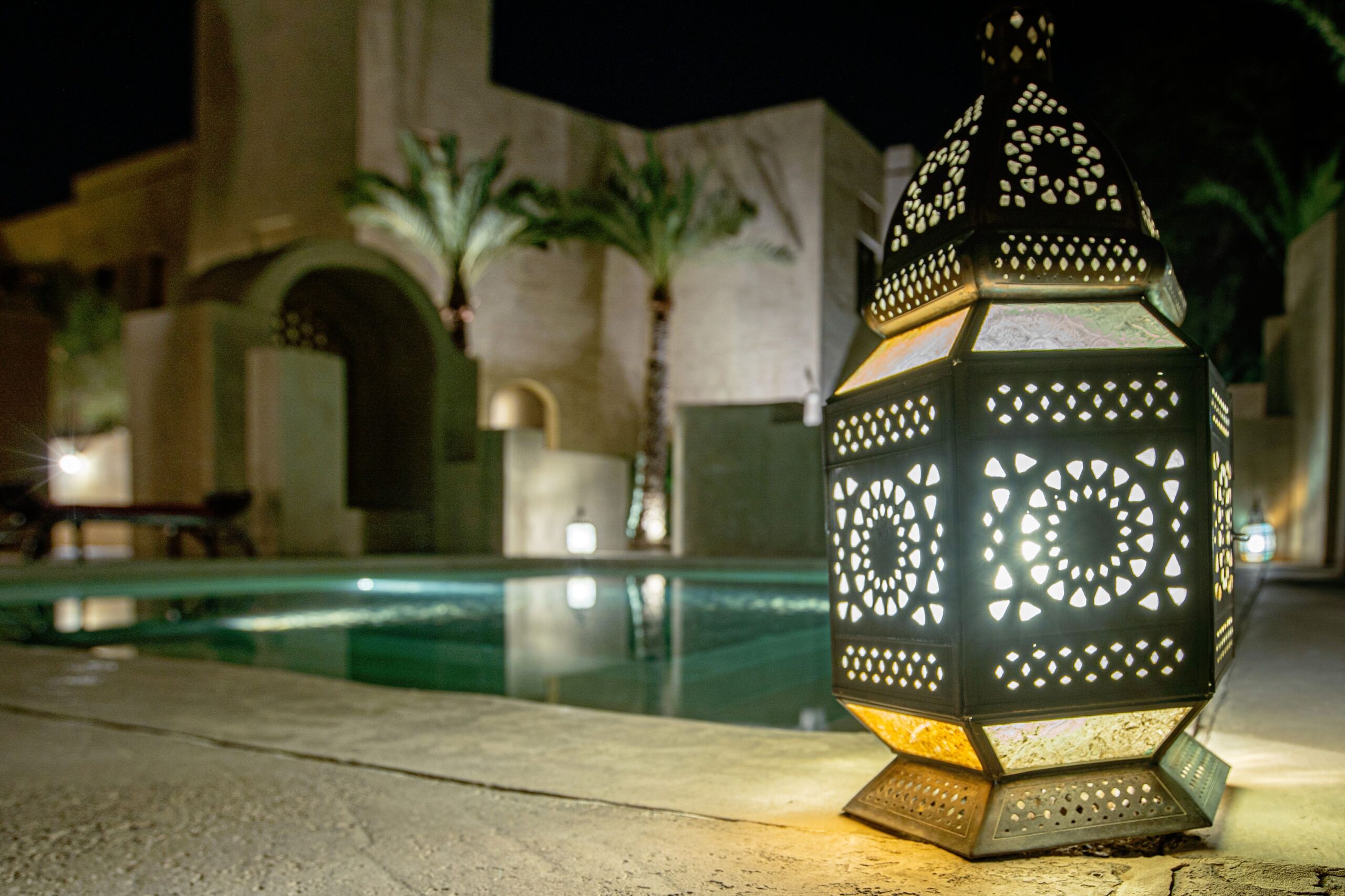 Ornate Andalusian-style lantern glowing beside a poolside at night with arched sandstone buildings