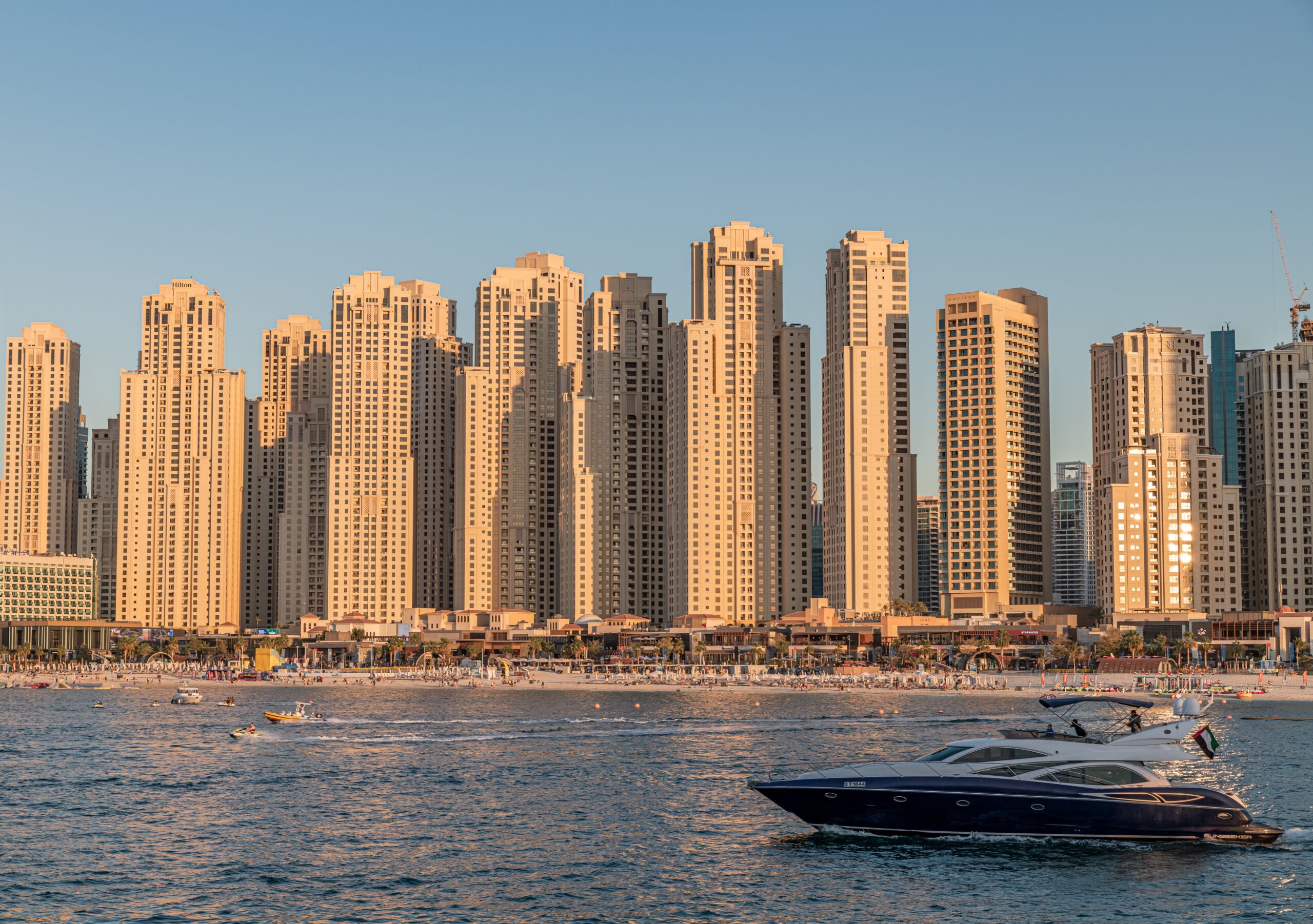 Golden-hour view of the Jumeirah Beach Residence (JBR) beachfront tower skyline from the water, with a motor yacht passing in the foreground, Dubai