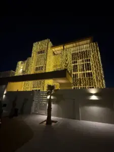 Front entrance of the villa at night, with string lights on the upper floors, a palm tree and a gate